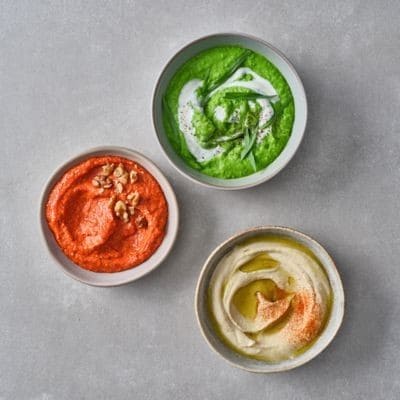Drei cremige Dips in Schalen: Hummus, ein roter Dip und ein grüner Dip auf grauem Hintergrund